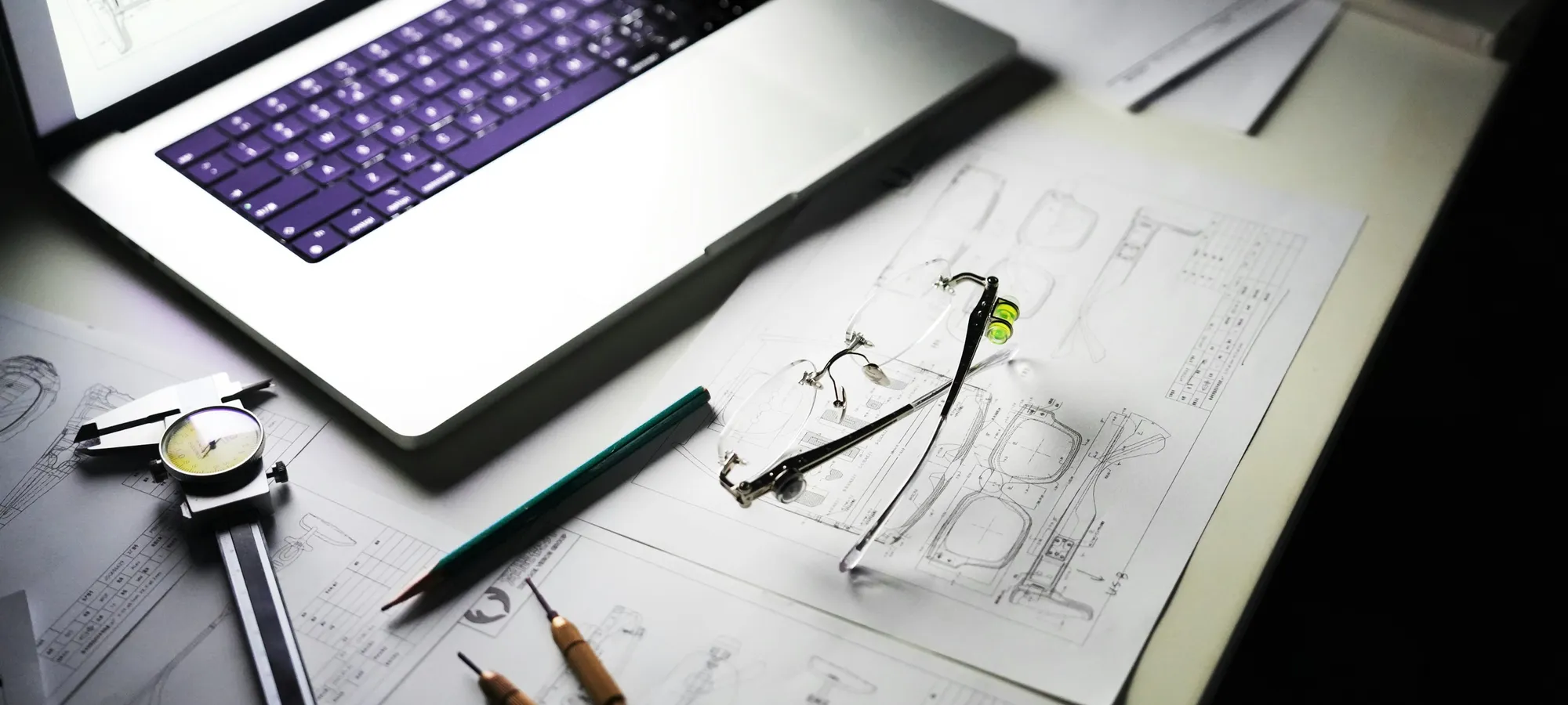 Architectural drawings, drafting pencils and calipers on a desk beside an open laptop