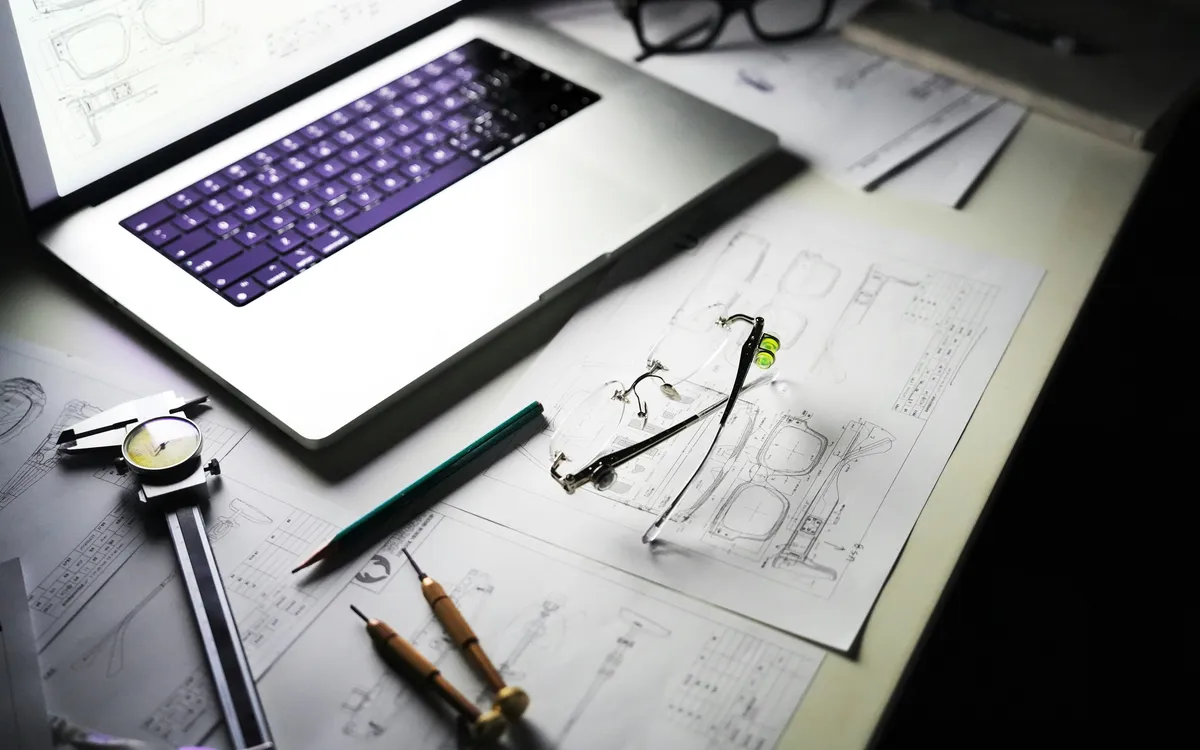 Architectural drawings, drafting pencils and calipers on a desk beside an open laptop